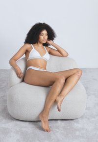 Iliektas, pūkains, gaiši pelēks krēsls ar teksturētu audumu. Modelis sēž, ģērbies baltā peldkostīmā, kājas sakrustotas, relaksēta poza. Mīksta paklāja fons.