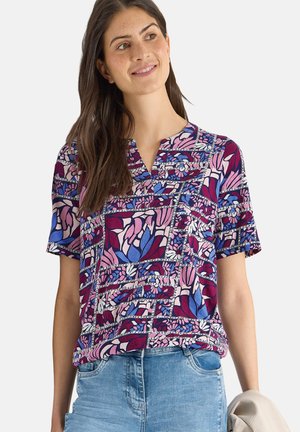 Femme portant un chemisier à manches courtes fleuri et coloré dans des tons de violet, rose et bleu, associé à un jean bleu clair, tenant une veste beige.