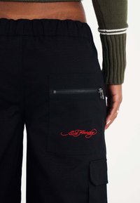Zwarte cargo broek met een ritszak en een rood geborduurd logo. De stof heeft een gestructureerde uitstraling en de tailleband is elastisch.