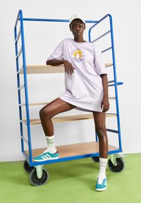 Vestido de camiseta oversized lila con un diseño gráfico de sol, combinado con zapatillas teal y calcetas a rayas, frente a una estantería azul y un suelo verde.