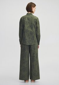 Olijfgroene bloemen shirt en brede broek set. Materiaal heeft geborduurde ontwerpen en subtiele texturen. Eenvoudige kraag en losse pasvorm.