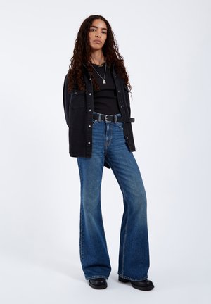 Young woman standing with curly hair, wearing black shirt, black button-up jacket, blue flared jeans, black belt, and black shoes.