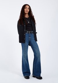 Black denim jacket, black top, and flared blue jeans. Curly hair, standing with hands behind. Minimalist silver necklace and belt.