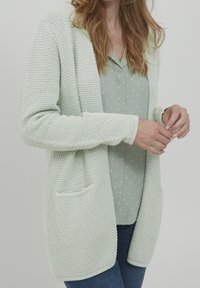 Cardigan di lana verde chiaro con un motivo testurizzato, con apertura frontale e due tasche. Abbinato a una camicetta verde chiaro a pois e jeans blu.