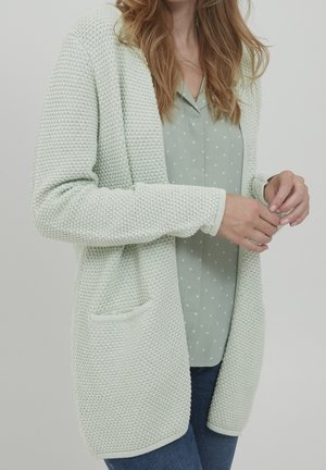 Lichtgroene, gebreide cardigan met een gestructureerd patroon, open voorkant en twee zakken. Gecombineerd met een lichtgroene blouse met stippen en blauwe jeans.