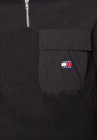 Schwarzer Strickpullover mit einem Reißverschlusskragen und einer Brusttasche. Die Tasche ist mit einem "Tommy Jeans"-Logo in rot, weiß und blau versehen.