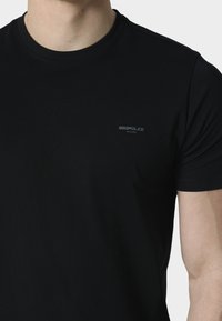 Uomo che indossa una maglietta nera semplice a girocollo con il logo "883POLICE MILANO" sul petto a sinistra, ritagliato sotto il mento e a metà braccio.