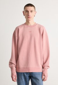 Sudadera rosa con cuello redondo y hombros caídos. Presenta un sutil logo "HUGO" en el centro. Textura suave y corte relajado.