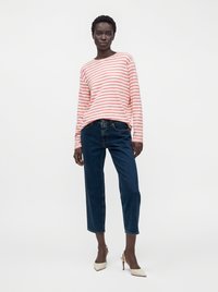 Femme debout portant une chemise à manches longues rayée rose et blanche, un jean court bleu foncé et des chaussures blanches à talons hauts sur un fond uni.