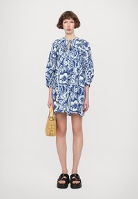 Blauwe en witte bloemenjurk met opgeblazen mouwen, een keyhole-halslijn en een losse pasvorm. Gecombineerd met een geweven gele tas en zwarte plateausandalen.