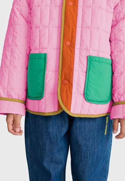 Enfant portant une veste matelassée rose avec des poches vertes et une patte de boutonnage orange, associée à un pantalon en jean bleu.