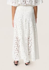 Weißer Spitzen-Maxi-Rock mit floralem Muster, transparenter Textur, einer definierten Taille und einem verdeckten Reißverschluss auf der Rückseite. Flache schwarze Absätze runden den Look ab.
