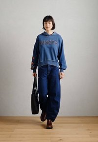 Μπλε φούτερ με κείμενο "HEROES", διακοσμημένο με λουλούδια στα μανίκια. Συνδυασμένο με τζιν παντελόνι φαρδιάς γραμμής και καφέ παπούτσια, κρατώντας μια μαύρη τσάντα.