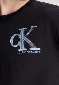 Černé bavlněné tričko s lehce modrým reflexním logem "cK" a nápisem "Calvin Klein Jeans" v bílé barvě umístěným na levé části hrudi. Krátký rukáv.