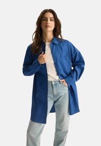 Kobieta z długimi brązowymi włosami, ubrana w niebieską oversize'ową koszulę na białej bluzce i jasnoniebieskich dżinsach, pozuje z jedną ręką w kieszeni.