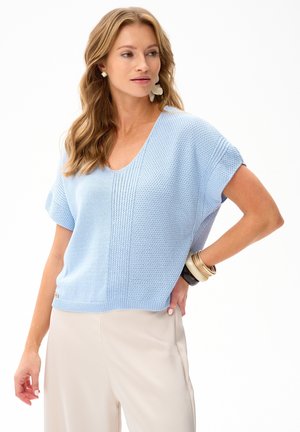 Kobieta z długimi blond włosami nosi jasnoniebieski dziergany top z krótkim rękawem i beżowe spodnie, uzupełnione kolczykami i wieloma bransoletkami.