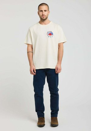 Homme debout face à l’objectif, portant un t-shirt beige à motif graphique, un pantalon bleu foncé et des chaussures marron devant un fond clair uni.