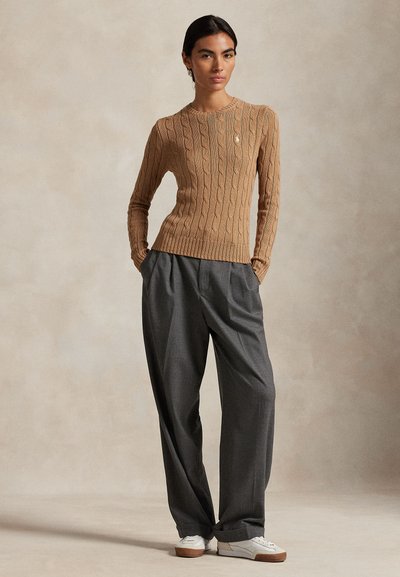 Kötött, világosbarna pulóver hosszú ujjakkal és bordázott szegéllyel, laza szürke nadrággal és fehér tornacipővel párosítva. Egyszerű, kerek bross díszítés.