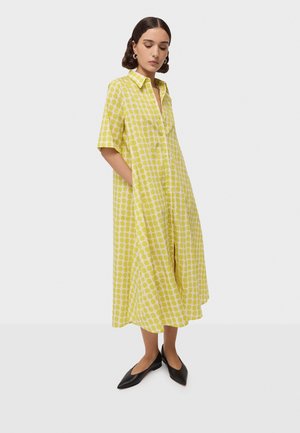 Femme portant une robe chemise midi jaune à pois blancs avec manches courtes et ballerines noires à bouts pointus, mains dans les poches, regardant vers le bas.