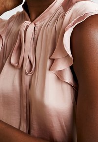 Silkesblus i mjuk rosa med volangärmar, nyckelhålsdetalj vid halsen och linjer. Slät textur och delikat design.