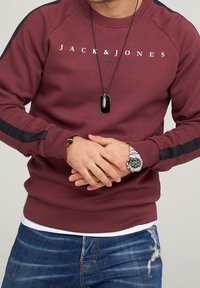 Man som bär en vinröd Jack & Jones sweatshirt, silverfärgad handledsklocka, svart hängsmycke och slitna blå jeans, med händerna ihopknäppta framför sig.