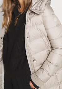 Zachte beige jas met capuchon, voorzien van een ritssluiting en drukknoopjes, gecombineerd met een gestructureerde zwarte trui en grijze gebreide manchetten.