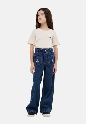 Jeune fille aux longs cheveux bruns portant un haut crème à manches courtes et un jean bleu taille haute avec des patchs d'ancre sur les poches avant.