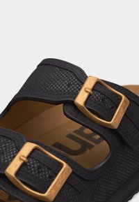 Μαύρο υφασμάτινο λουρί σανδαλιού με δύο μπρονζέ αγκράφες πάνω σε ταμπα πέλμα που δείχνει μερικό κείμενο εμπορικού σήματος στο εσωτερικό.