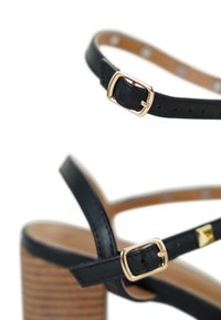 Zwarte leren hoge sandalen met hak, voorzien van een gouden gespsluiting, met een gestructureerde bruine houten blokhak en kruislings verlopende banden.