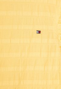 Żółta fakturowana tkanina z subtelnymi poziomymi paskami oraz małym wyszywanym logo Tommy Hilfiger w kolorze czerwonym, białym i granatowym.