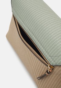 Gros plan sur un sac beige texturé et vert pâle avec une fermeture éclair dorée partiellement ouverte et une languette en cuir brun.