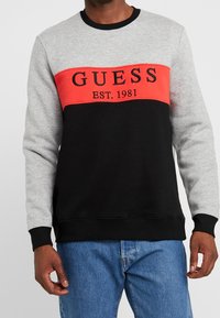 Sudadera bloqueada en gris, negro y rojo. Presenta el logo "GUESS" y el detalle "EST. 1981" en el pecho. Puños y cuello acanalados. Tejido suave.