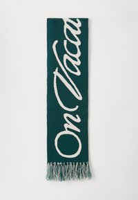 Cachecol em teal com texto em creme tricotado "Em Férias". Apresenta franjas nas extremidades e um acabamento macio e texturizado.