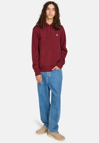 Hoodie bourgogne avec une poche avant et un cordon de serrage, associé à un jean bleu clair ample et des chaussures beiges, dotées d'un tissu texturé.