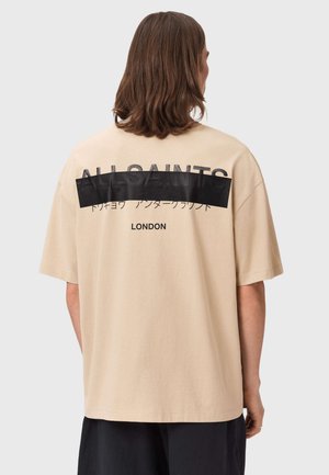 Osoba o brązowych włosach do ramion, ubrana w beżowy T-shirt z czarnym napisem i grafiką na plecach oraz czarne spodnie.