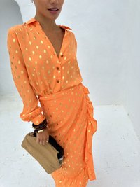 Oranger Kleid mit goldenen Punkten, Knopfleiste vorne, Kragen, rüschenseitlich. Getragen mit einer beigen Clutch und dunklen Holzarmreifen.
