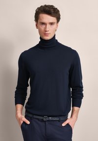 Navyfarbener Rollkragenpullover aus glattem Strickstoff. Enganliegende Silhouette mit langen Ärmeln und gerippten Bündchen, kombiniert mit dunklen Hosen und einem Gürtel.