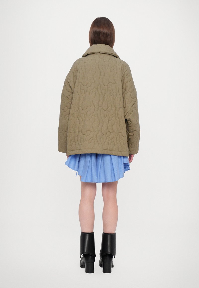 Veste matelassée olive avec un grand col et des coutures abstraites, associée à une jupes plissée bleu clair et des bottes à talons épais noires.