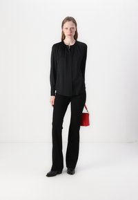 Blusa nera a maniche lunghe con scollo arricciato, abbinata a pantaloni neri a palazzo e stivali neri con tacco, completata da una borsa rossa.