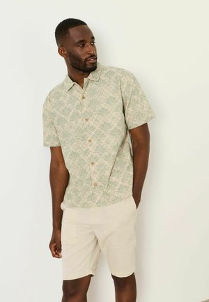 Mann trägt ein kurzärmliges Hemd mit einem grünen Palmenblattmuster und hellbeige Shorts, steht mit einer Hand in der Tasche.