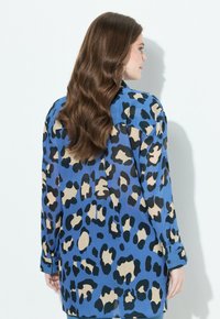 Donna con lunghe onde di capelli castani, che indossa una camicia a maniche lunghe blu con stampa leopardata, in piedi contro uno sfondo bianco, rivolta di lato.