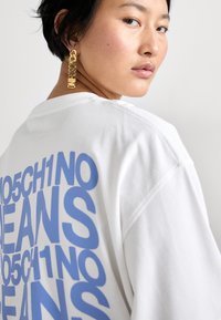 Balta hoodie ar lieliem ziliem burtiem, uz kuriem rakstīts "MOSCHINO JEANS", un uz aizmugures "105CH1NO". Modelis nēsā zelta auskarus.