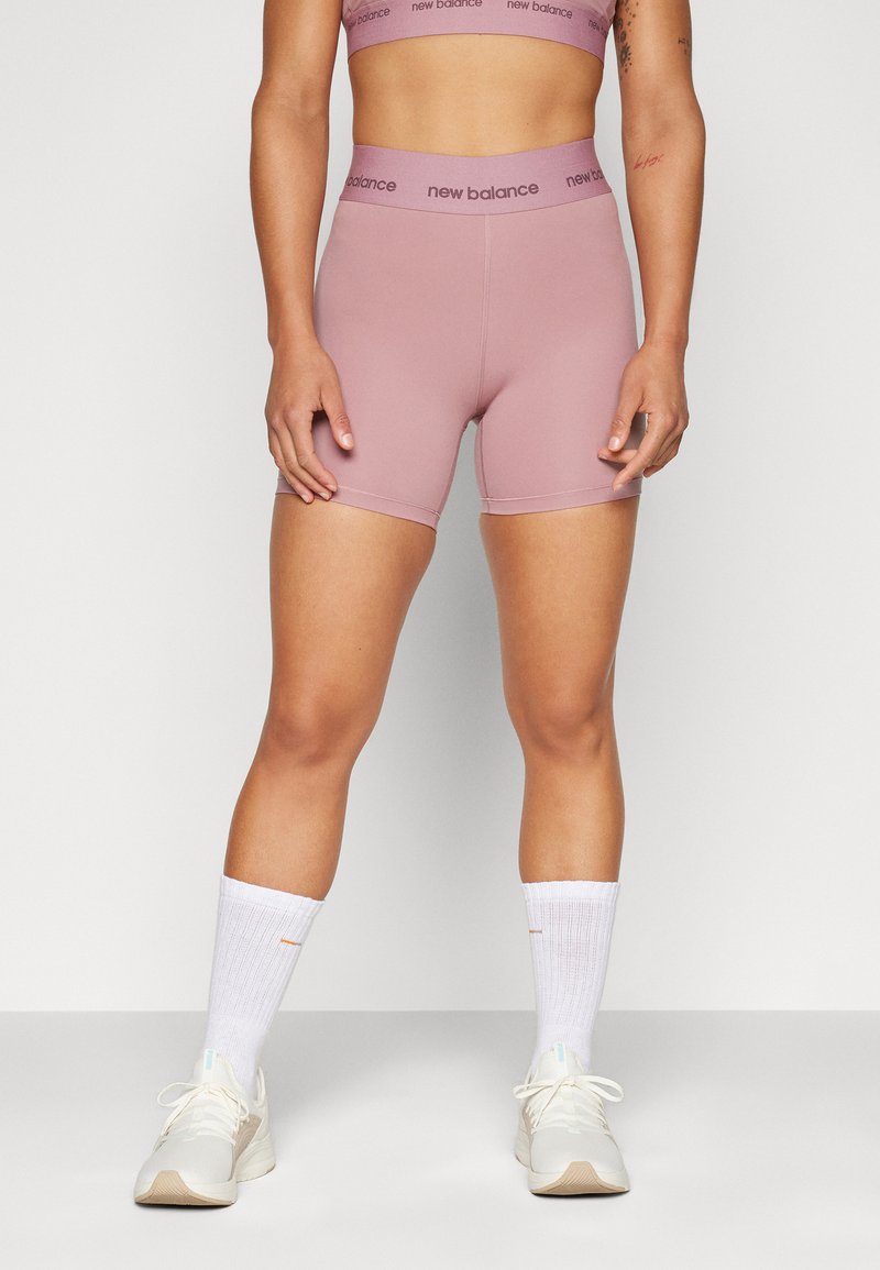 Pessoa em forma vestindo calções de atletismo mauve da New Balance, soutien desportivo a condizer, meias brancas até ao tornozelo e sapatilhas beiges, de pé contra um fundo liso.