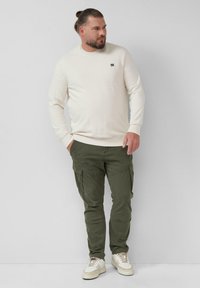 Man met baard en knot die een crèmekleurige sweatshirt, olijfkleurige cargobroek en witte sneakers draagt, staand tegen een effen lichte achtergrond.