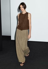 Zandkleurige suede mouwloze top met een diagonale knopenlijst, gecombineerd met losse olijfgroene broek en eenvoudige bruine sandalen.