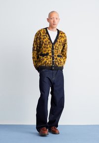 Lepardmønstret cardigan i gul og sort, strikket med en blød tekstur, featuring en V-hals, frontknapper og to lommer. Mørke denim bukser.