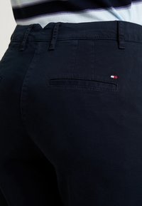 Granatowe spodnie o gładkiej fakturze, z jedną tylną kieszenią i małym logo w kolorach czerwonym, białym i niebieskim.