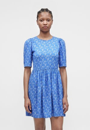 Frau mit geflochtenem Haar, die ein blaues Blumenkleid mit Puffärmeln und geraffter Taille trägt, steht vor einem einfarbigen hellen Hintergrund.