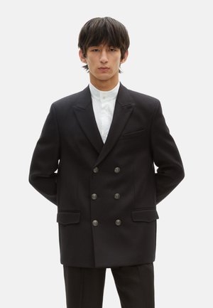 Blazer negro de doble botonadura hecho de tela suave, con botones plateados, dos bolsillos frontales y un diseño de solapa muesca. Camisa blanca debajo.