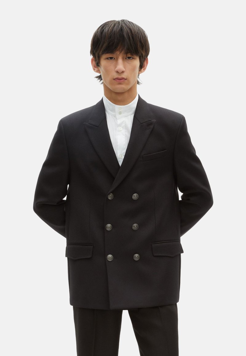 Schwarzes, doppelt geknöpftes Blazer aus glattem Stoff, mit silbernen Knöpfen, zwei vorderen Taschen und einem Reverskragen. Weißes Hemd darunter.